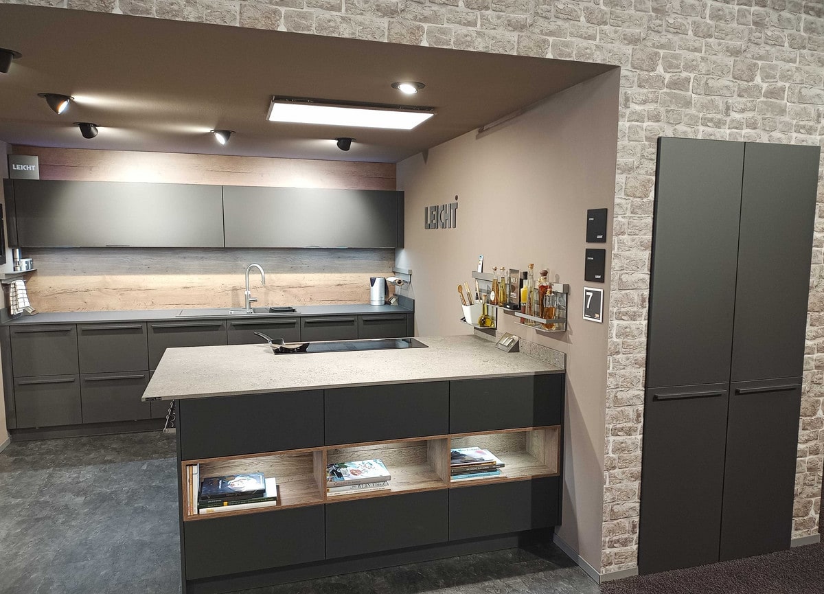 Dunkle matte Küche in einer Nische der Steinwand: Rückwand in Eichenoptik, Halbinsel mit heller Keramikplatte und beleuchteten offenen Fächern mit Kochbüchern, rechts ein hoher Schrank