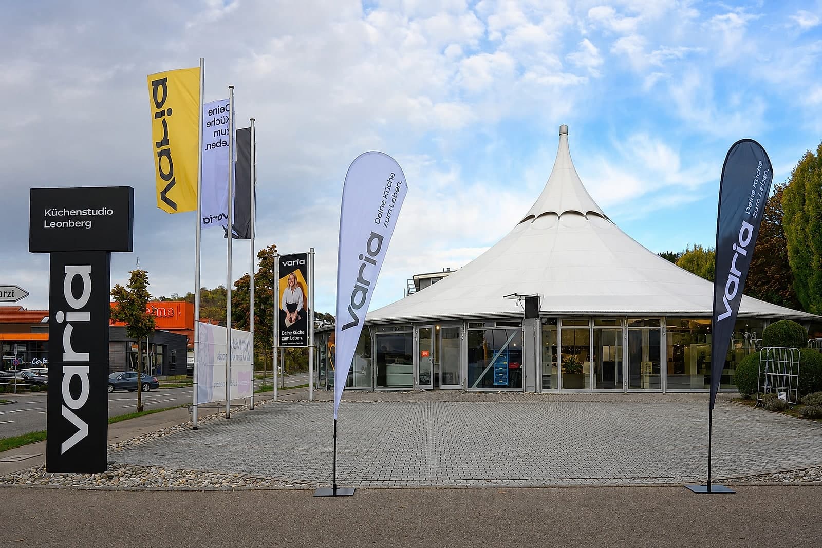 Der Pavillon in Leonberg von der Straße aus im Oktober 2026: Pylon „Küchenstudio Leonberg“, Fahnen und Beachflags von varia, dahinter die Glasfront unter dem weißen Dach