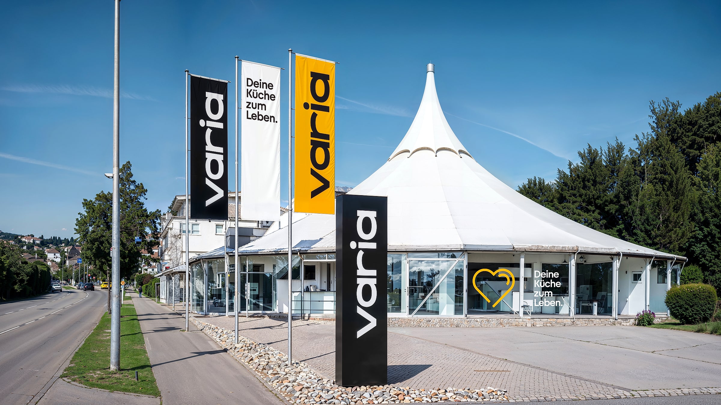 Visualisierung des Pavillons in Leonberg im neuen Auftritt: varia-Fahnen, schwarzer Pylon und ein gelbes Herz an der Glasfront unter dem weißen Dach, blauer Himmel