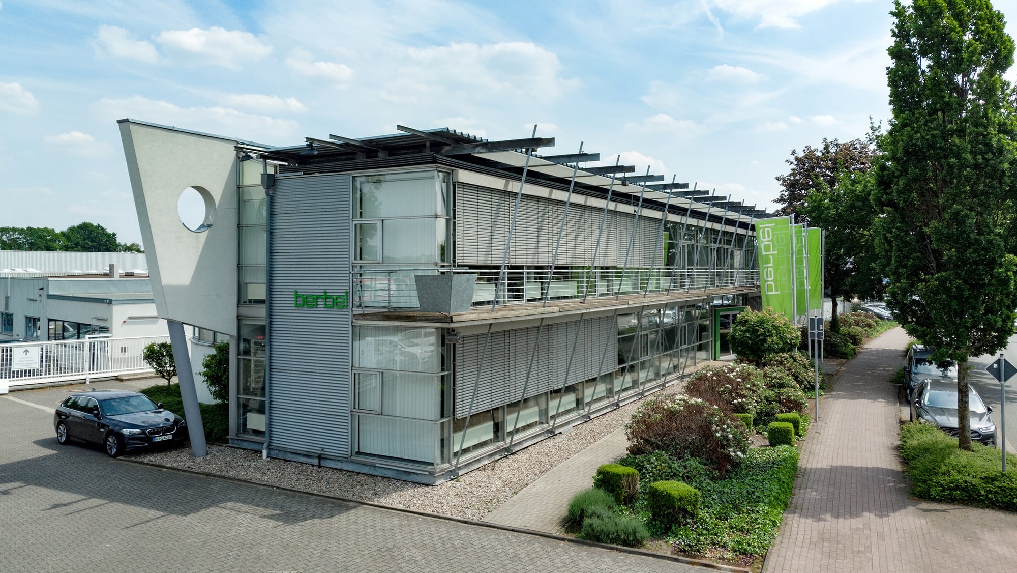 Firmengebäude von berbel: ein zweigeschossiger Bau mit Glasfassade und Sonnenschutzlamellen, davor eine bepflanzte Einfahrt