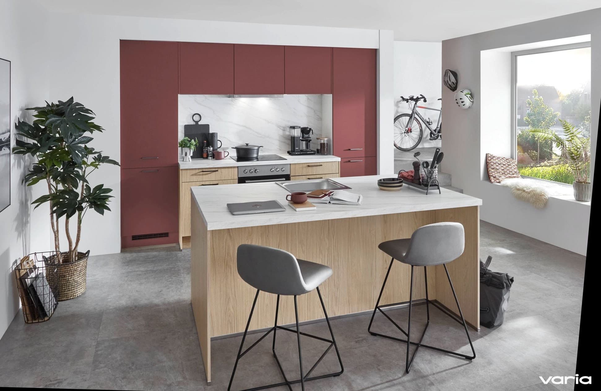 Moderne Küche mit rostroten Hochschränken und einer Kochinsel in heller Eiche mit weißer Steinoptik-Platte, darauf Laptop, aufgeschlagenes Buch und Tasse, davor zwei graue Barhocker, im Hintergrund ein Rennrad an der Wand und eine Fensterbank mit Fell und Kissen