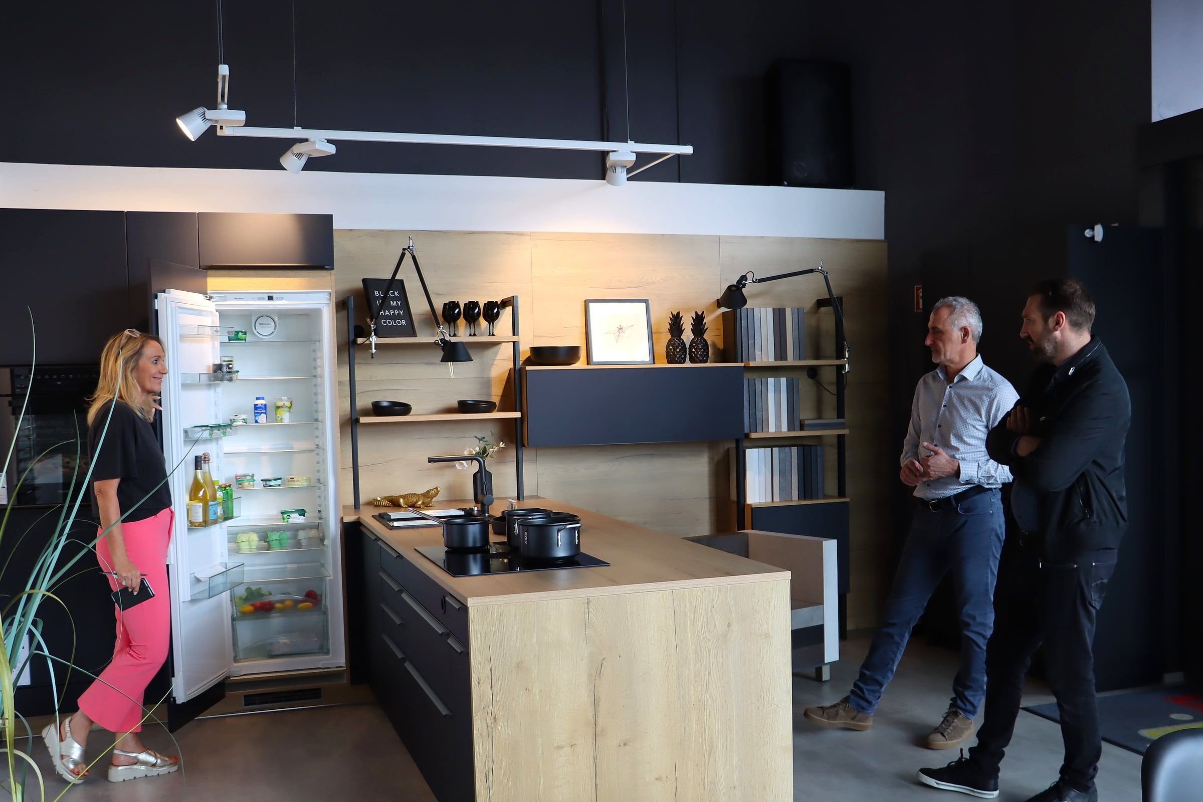 Ausstellungsküche in Emmendingen: Kochinsel in Eiche mit schwarzen Fronten und Kochfeld, dahinter offene Regale vor einer Holzwand und ein geöffneter Kühlschrank im Hochschrank; Boris Scholl spricht mit zwei Besuchern