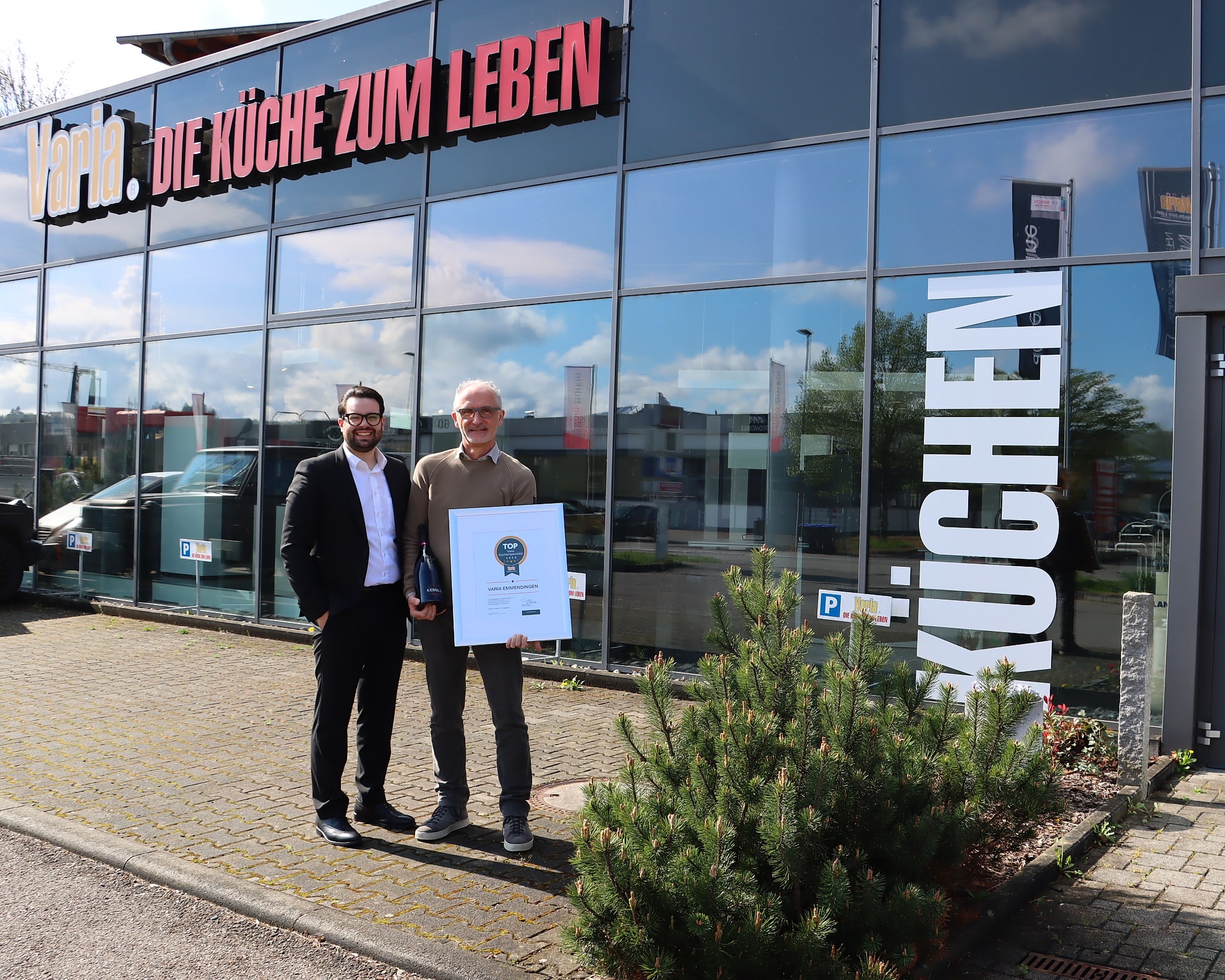 Boris Scholl vor seinem Küchenstudio am Elzdamm in Emmendingen, in der Hand die Urkunde zur Nominierung als TOP Küchenspezialist 2025, neben ihm ein Kollege; dahinter die lange Glasfront mit dem alten Schriftzug Varia Die Küche zum Leben und dem Wort Küchen auf den Scheiben