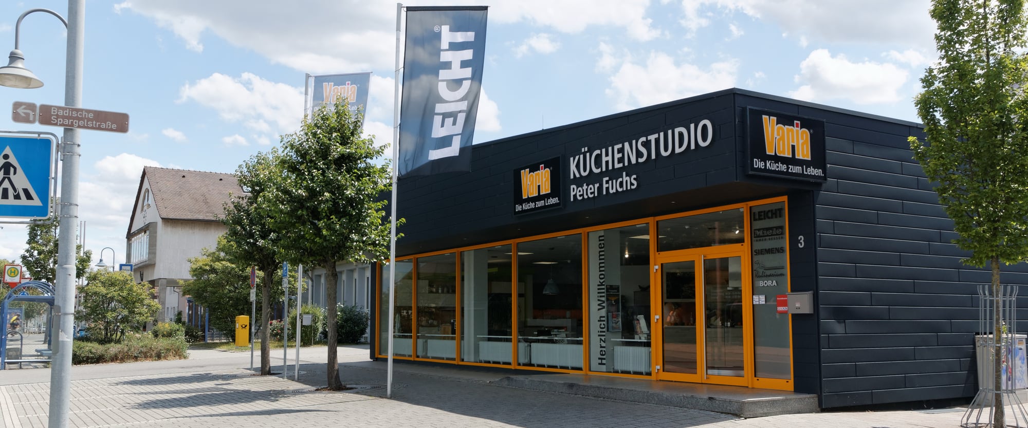 varia Küchenstudio Karlsdorf von der Straße aus gesehen: dunkle Fassade mit Varia-Schriftzug, orange gerahmte Schaufenster und Eingang unter blauem Himmel