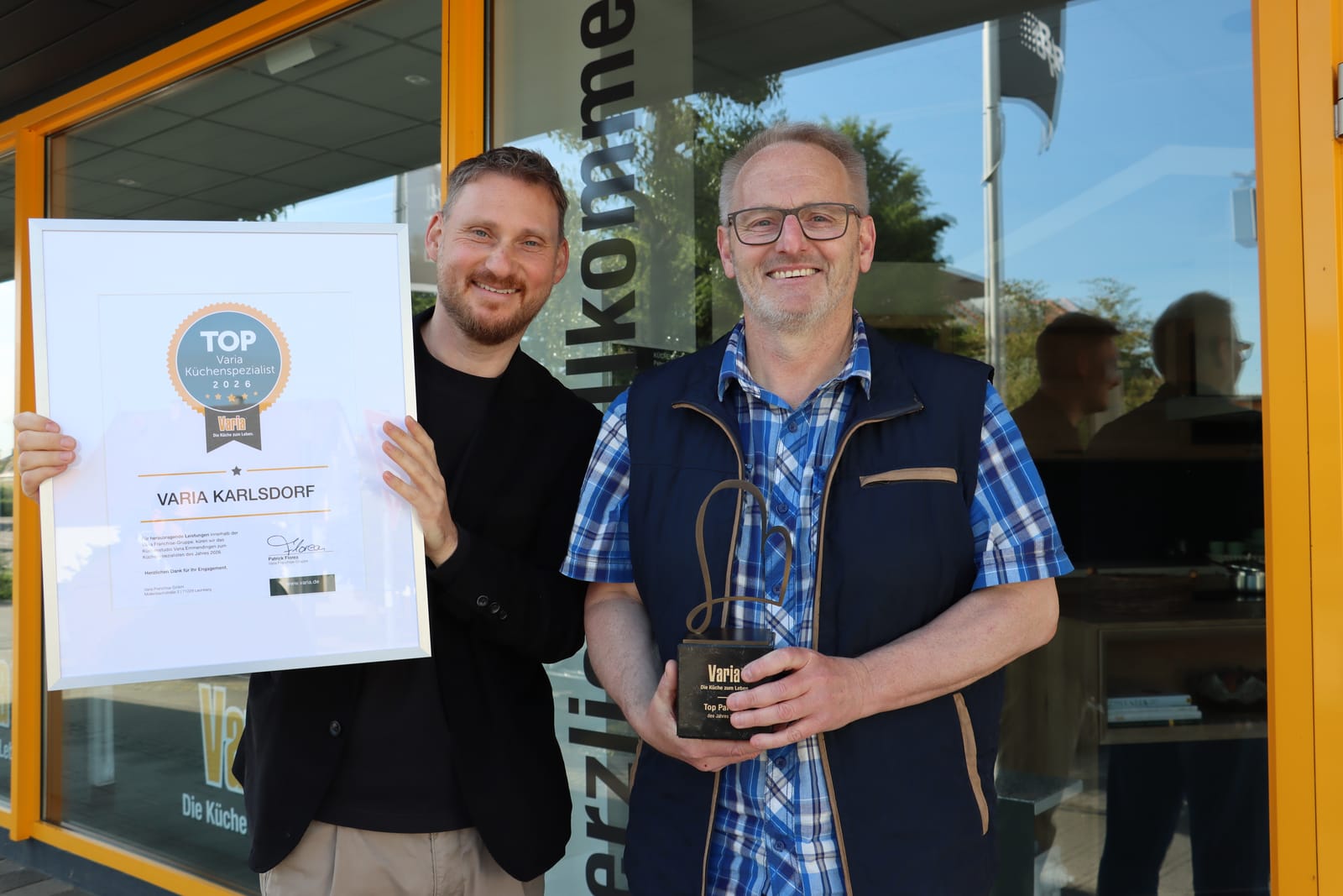 Peter Fuchs vor dem Studio in der Amalienstraße mit dem Pokal „Top Partner des Jahres“, daneben Patrick Florea von varia mit der Urkunde „TOP varia Küchenspezialist 2026“
