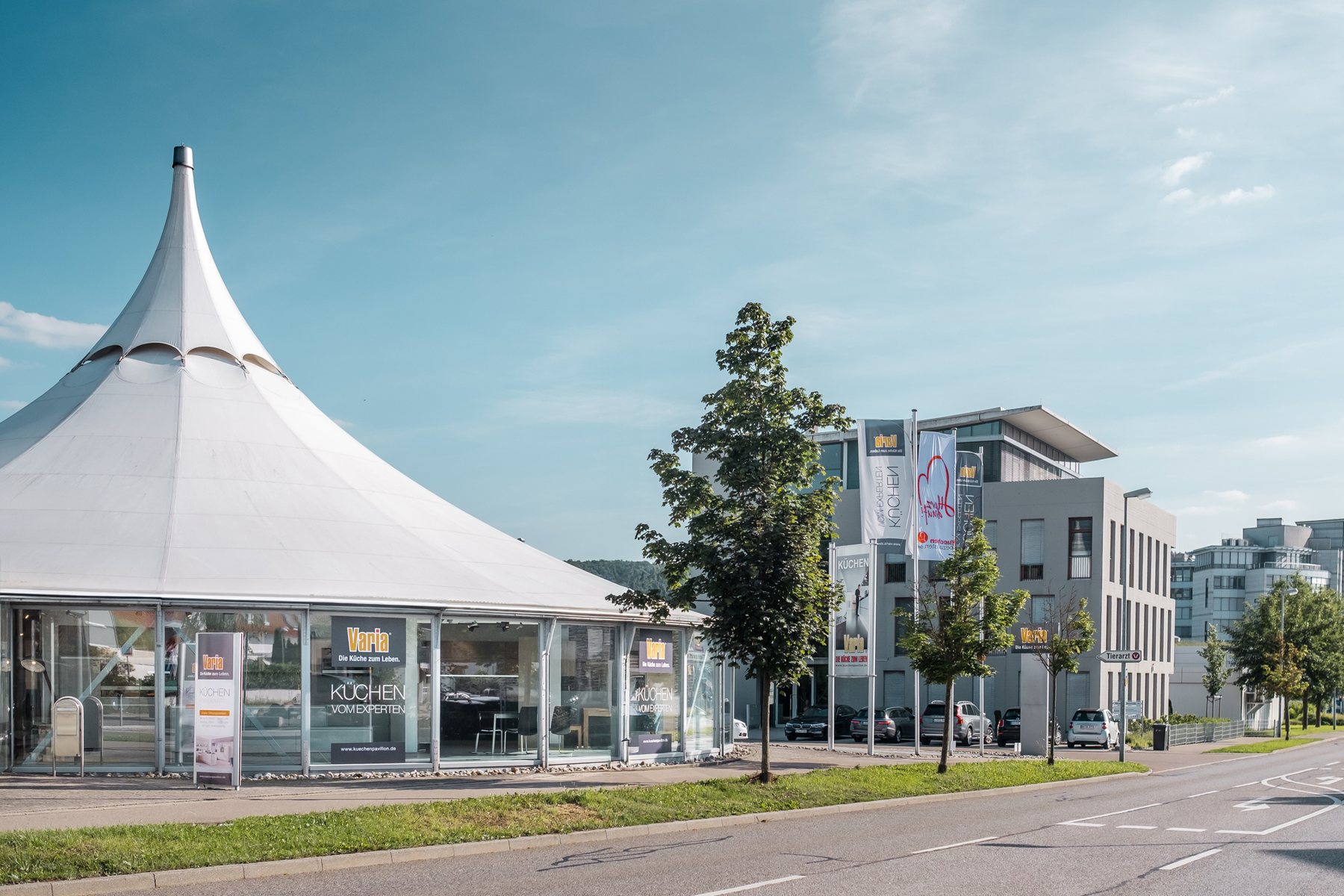 Der Frei-Otto-Pavillon in Leonberg von der Straße aus: das weiße Dach über der umlaufenden Glasfront, rechts die Fahnen, blauer Himmel
