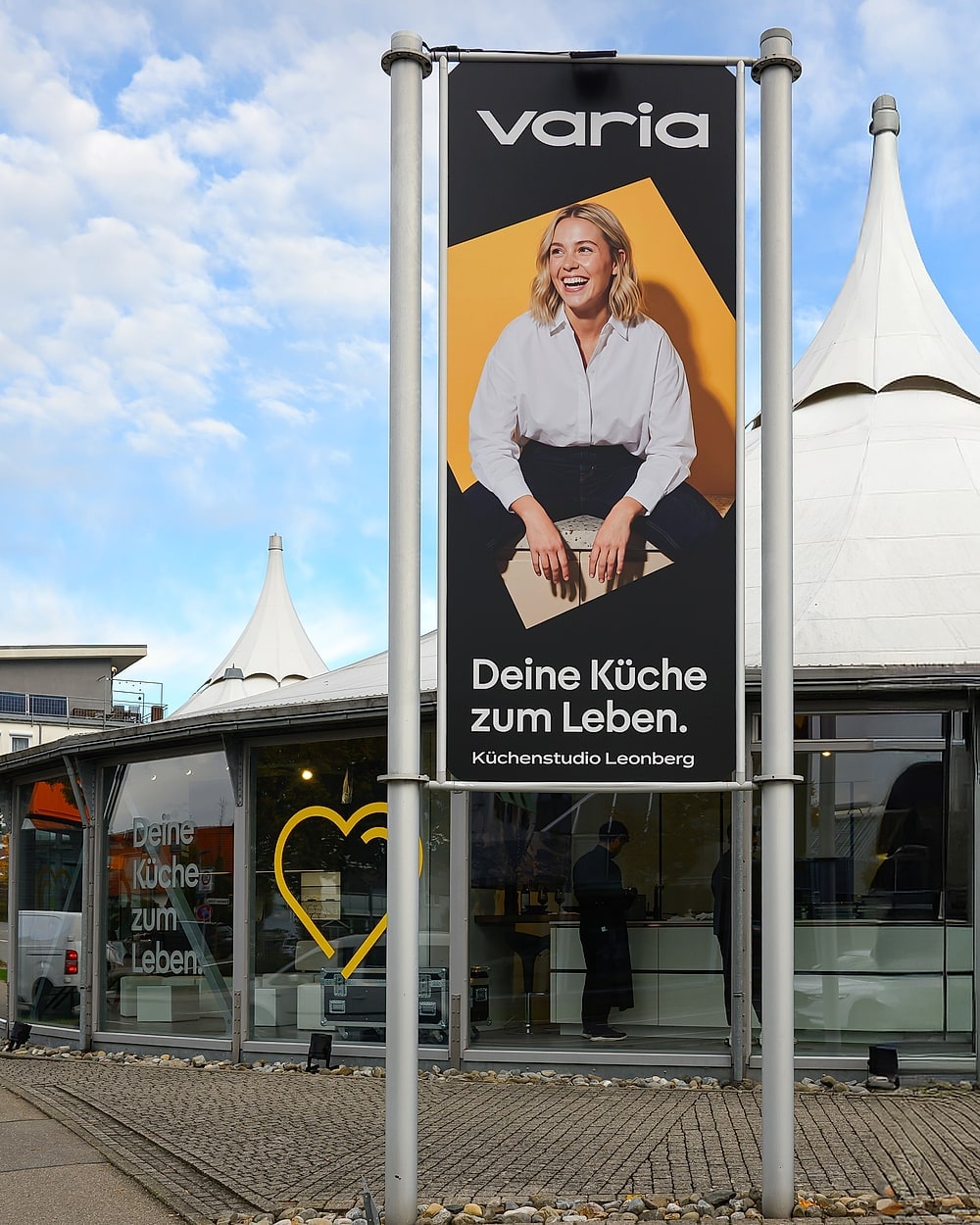 Banner von varia vor dem Pavillon: „Deine Küche zum Leben. Küchenstudio Leonberg“, dahinter die beiden weißen Dachspitzen und das gelbe Herz an der Glasfront