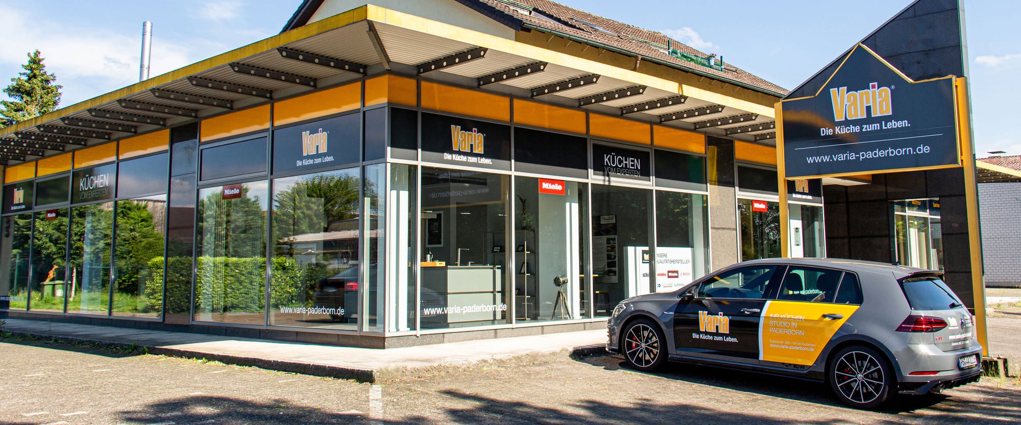 Das Küchenstudio in Paderborn von der Straße aus: umlaufende Schaufensterfront unter dem gelben Vordach, rechts der Pylon, davor der Studiowagen
