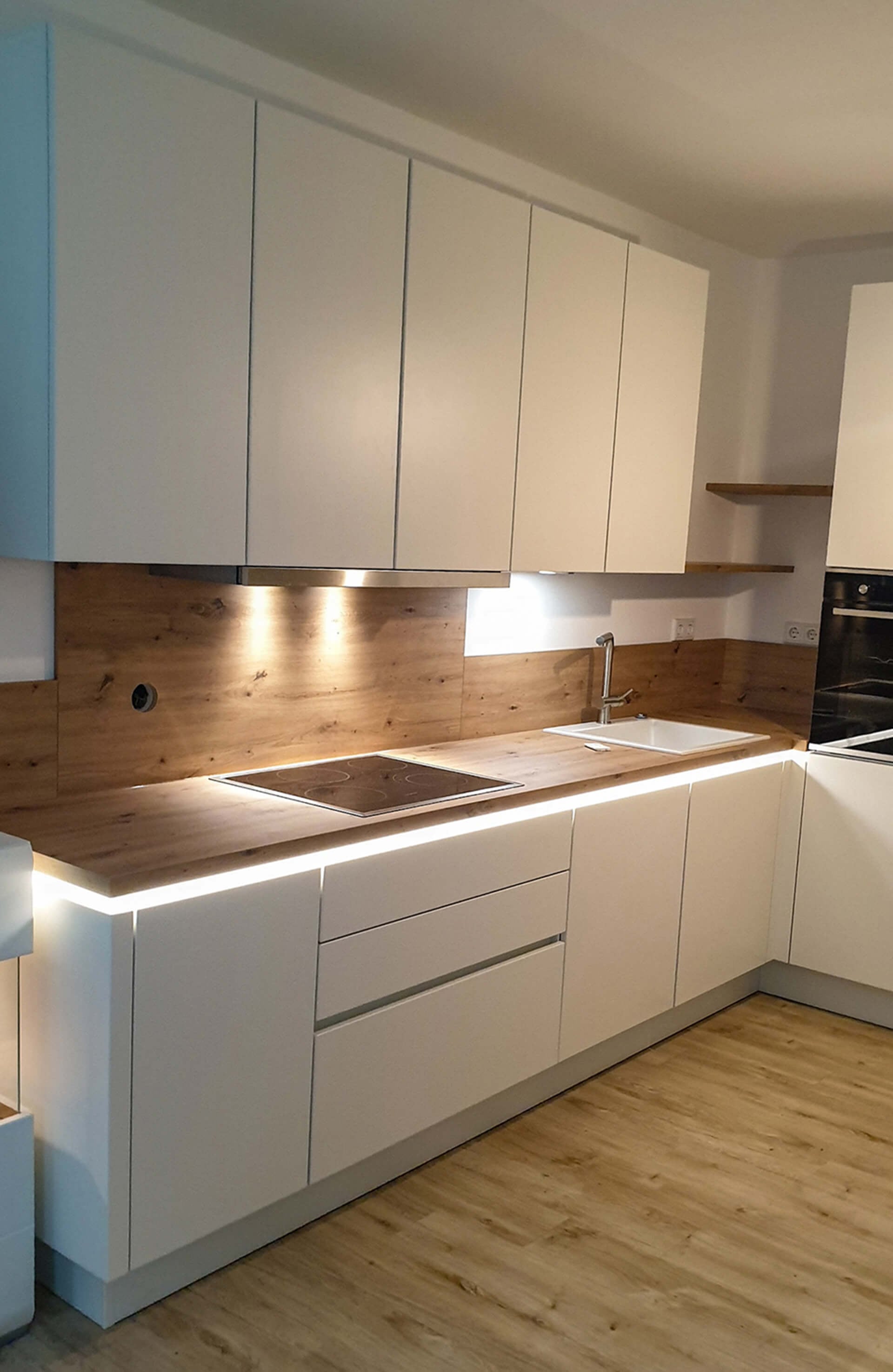 Fertig montierte Kundenküche: weiße grifflose Fronten, Arbeitsplatte und Rückwand in Eichenoptik, Lichtleiste unter der Platte, Holzboden