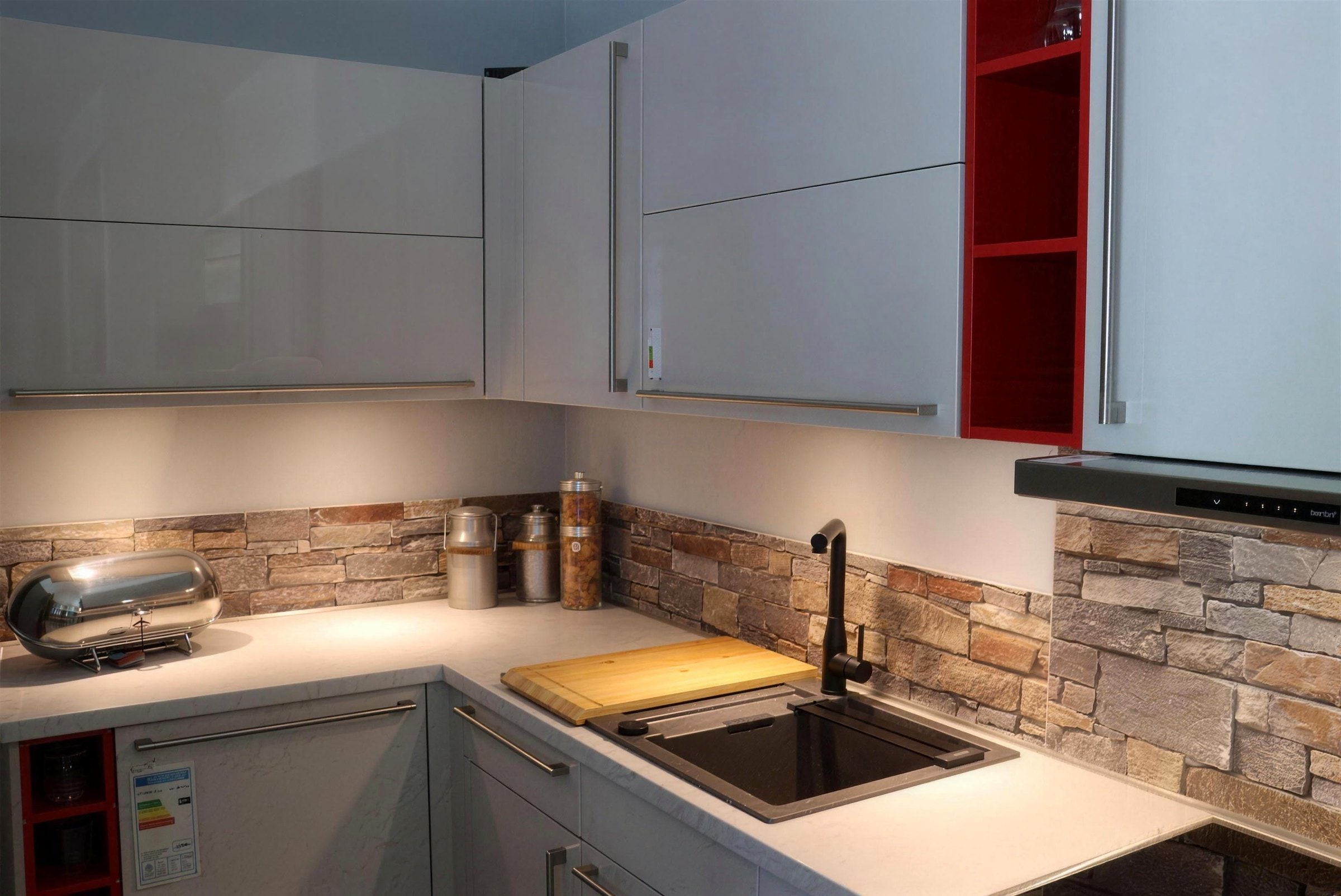 Purion-Eckküche in Grau Hochglanz mit Rückwand in Steinoptik, weißer Arbeitsplatte, Spüle in Schwarz mit Holzbrett und einem roten offenen Regal