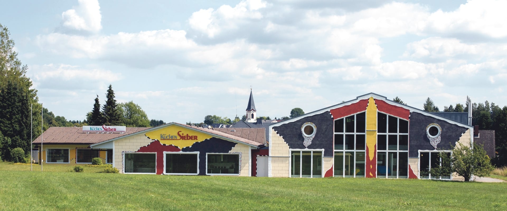 Küchenstudio in Weißdorf von außen: langgestrecktes Gebäude mit farbig gestalteter Fassade, runden Fenstern, großen Schaufenstern und Wiese davor