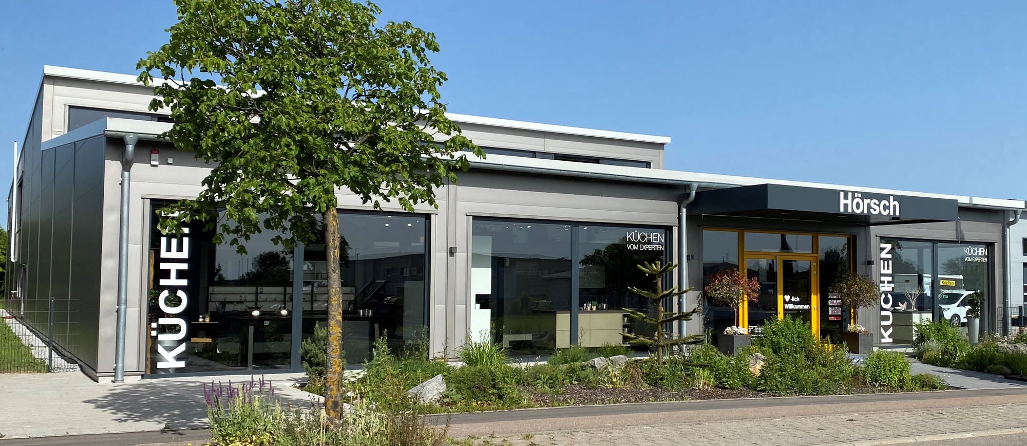 Das Küchenstudio in Welzheim von der Straße aus: grau verkleideter Neubau mit großen Schaufenstern, dem Wort KÜCHEN in großen weißen Buchstaben und einem gelb gerahmten Eingang, davor ein junger Baum