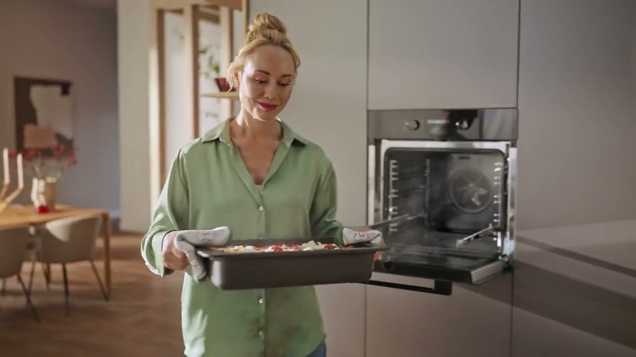 Frau trägt ein volles Blech vom hoch eingebauten Backofen zur Arbeitsplatte und stellt es ab