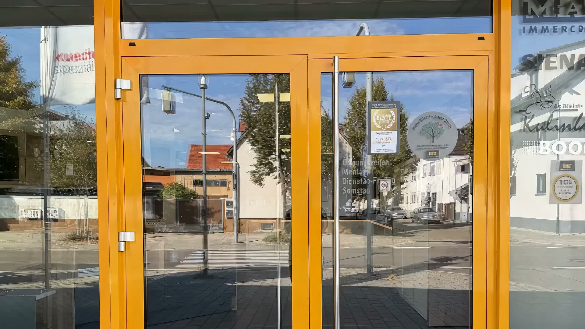 Rundgang durch das Küchenstudio in Karlsdorf: durch die orange gerahmte Glastür hinein, an der weißen Küchenzeile und der Musterwand entlang, vorbei an der Kochinsel unter der grünen Decke und am runden Tisch bis zur dunklen Küche an der blauen Wand