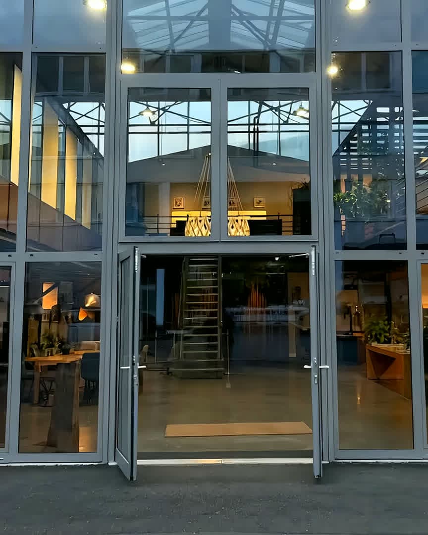 Flug durch das KüchenLoft in Köln: von der hohen Glasfront durch die offene Tür in die Halle, über den langen Tisch und an der Stahltreppe vorbei hinauf zur großen Leuchte unter dem Glasdach, dann ein Regal mit farbigen Musterfronten, Hochschränke in Nussbaum hinter einer dunklen Steininsel und zum Schluss eine helle Küche mit Eichenkanten und dem Blick zur Küche in Terracotta