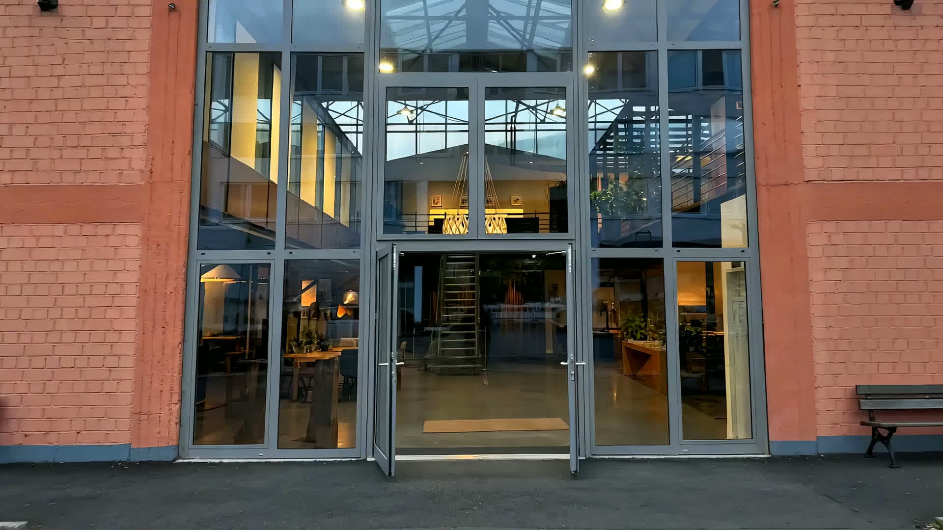 Flug durch das KüchenLoft in Köln: von der hohen Glasfront durch die offene Tür in die Halle, über den langen Tisch und an der Stahltreppe vorbei hinauf zur großen Leuchte unter dem Glasdach, dann ein Regal mit farbigen Musterfronten, Hochschränke in Nussbaum hinter einer dunklen Steininsel und zum Schluss eine helle Küche mit Eichenkanten und dem Blick zur Küche in Terracotta