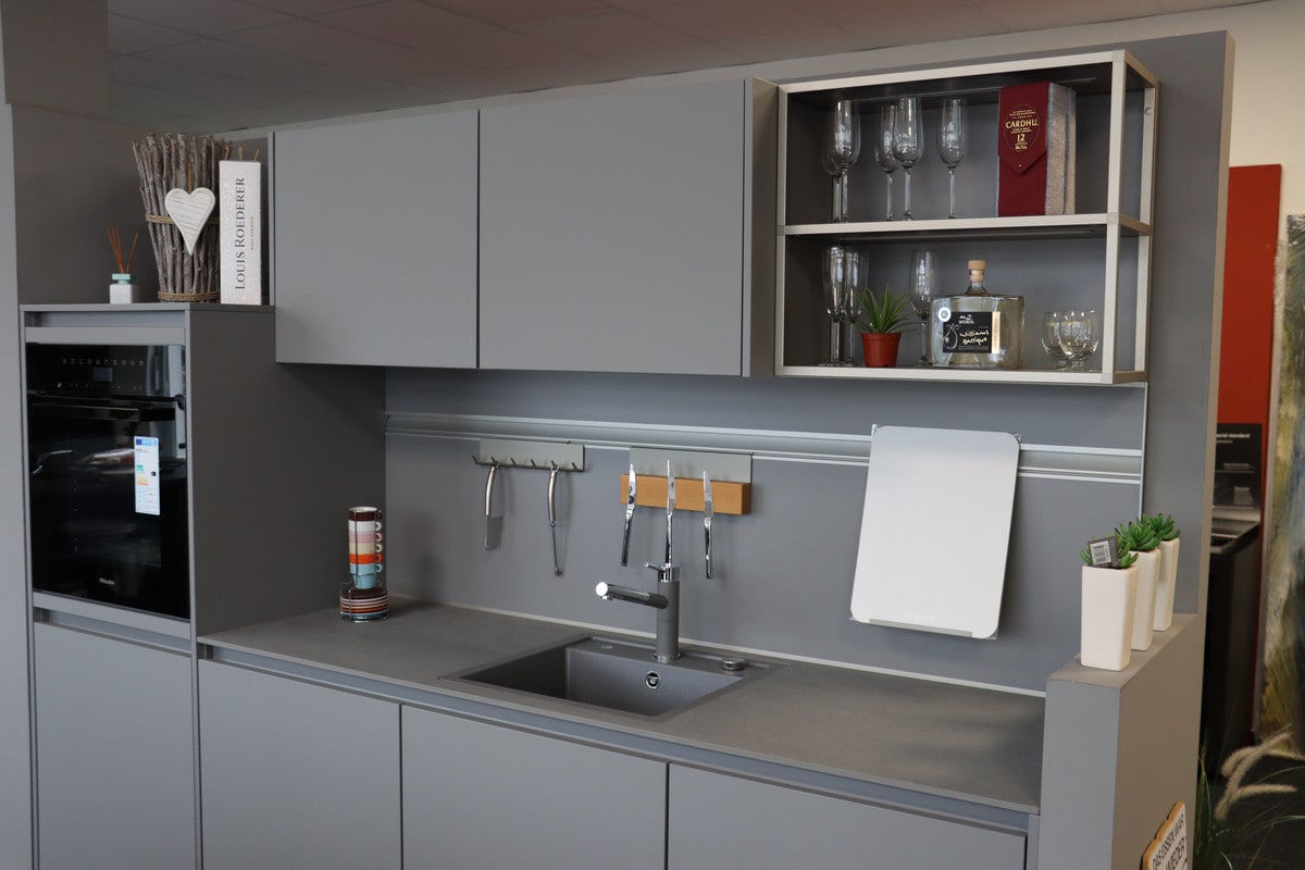 Schüller-Küchenzeile in Mattgrau ohne Griffe: Hochschrank mit Backofen, Spüle in Dunkelgrau, darüber eine Reling mit Messern und ein offenes Regal in Aluminiumoptik mit Gläsern