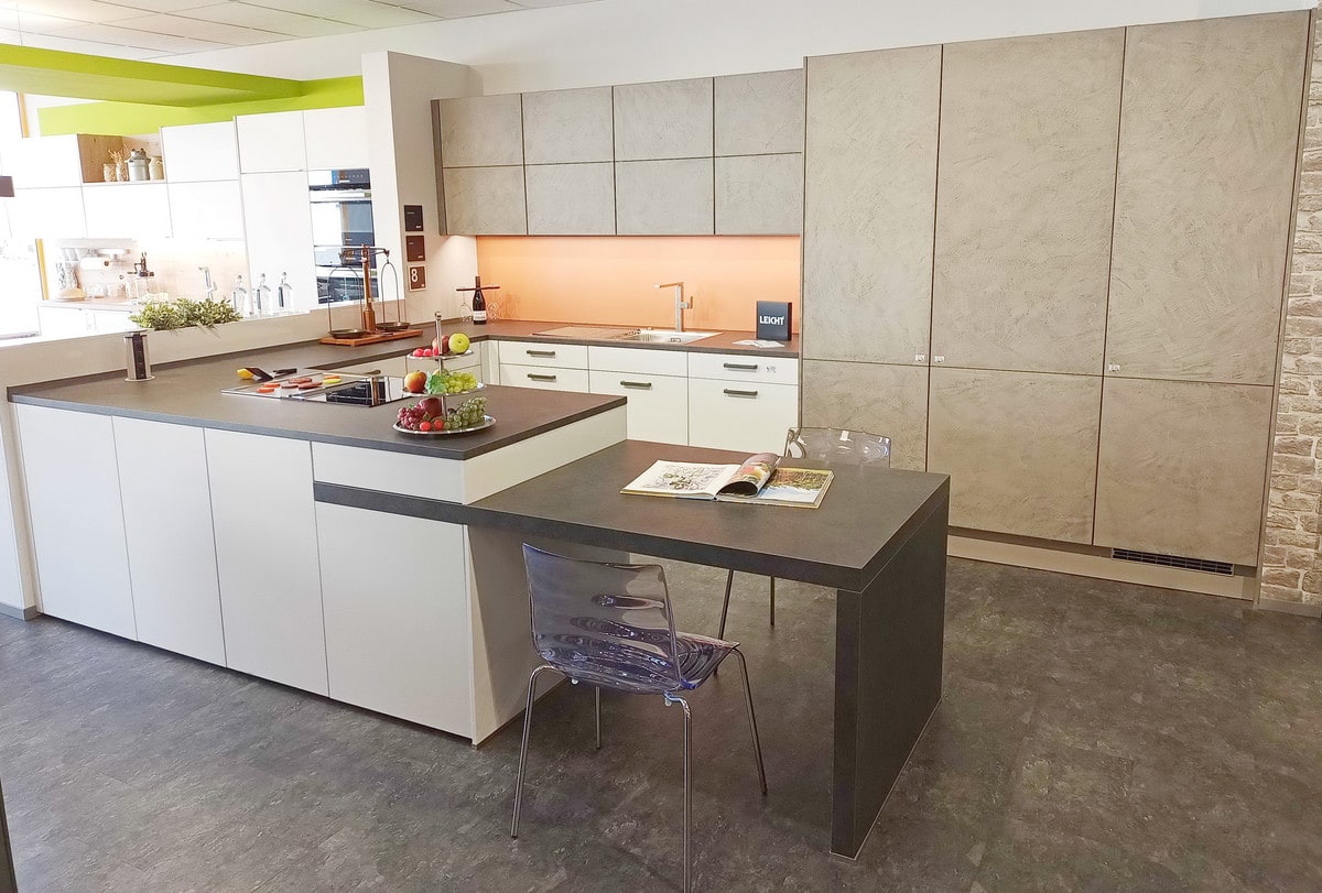 Ausstellungsküche in Karlsdorf: cremeweiße grifflose Kochhalbinsel mit dunkler Arbeitsplatte und angesetztem Tisch, dahinter hohe Schränke mit Betonfronten in Spachteltechnik und eine terrakottafarbene Nischenwand
