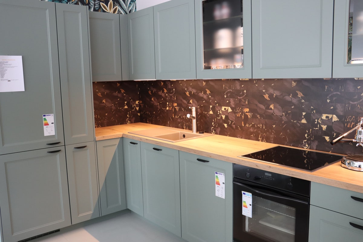 Landhausküche in Salbeigrün mit Rahmenfronten und schwarzen Griffen, Arbeitsplatte in heller Holzoptik, Spüle in Sandfarbe und dunkle Rückwand mit goldenen Flecken